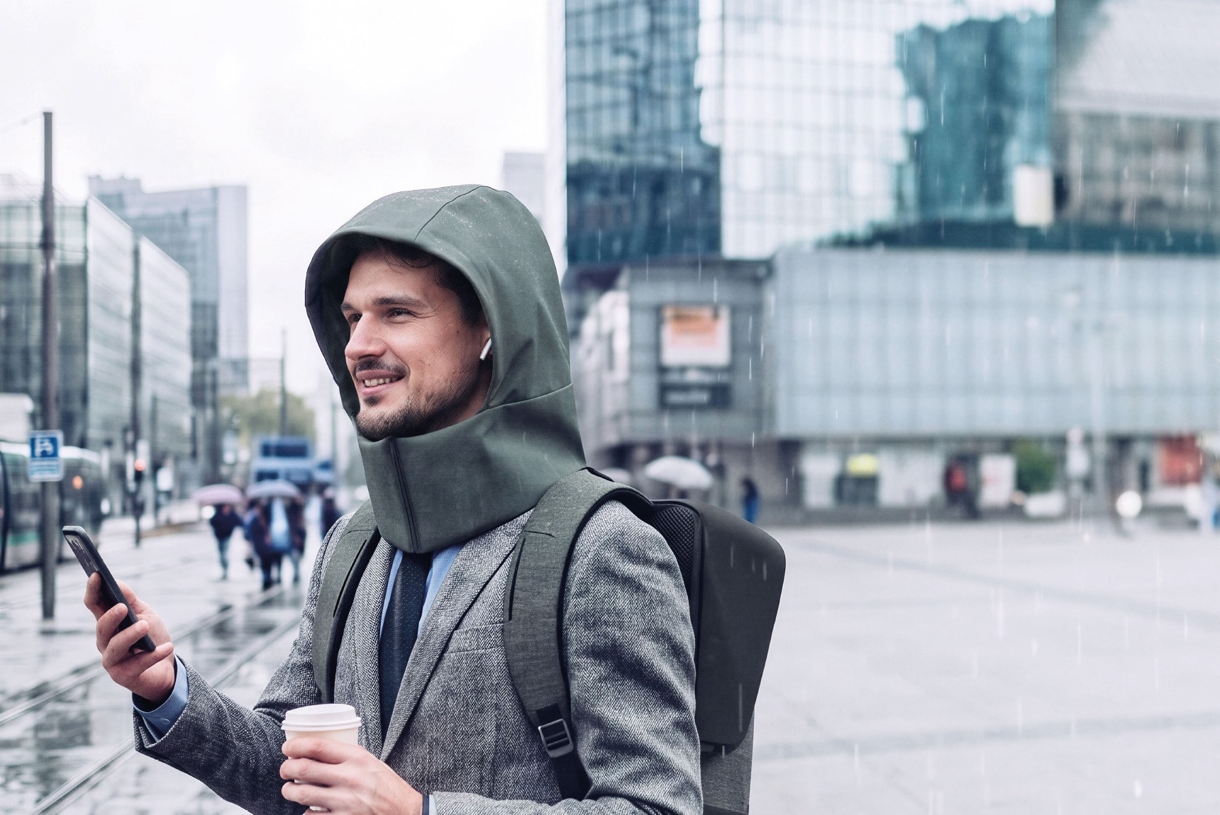 Un homme sous la pluie en ville, la capuche BAGRAIN déployée sur la tête, téléphone et café en main