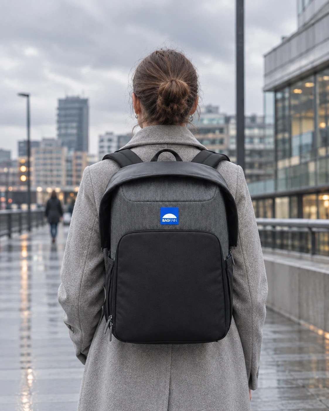 Une femme vue de dos sur une passerelle mouillée en ville, le sac BAGRAIN sur les épaules, logo bleu bien visible et capuche repliée